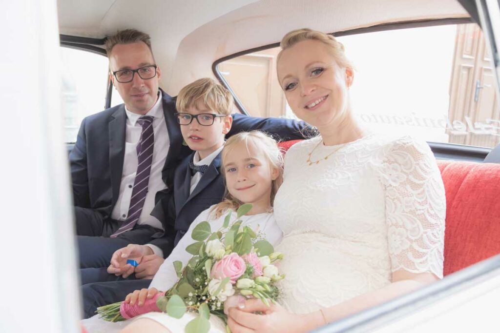 Brautpaar sitzt mit seinen Kindern in einem Auto und schauen in die Kamera des Hochzeitsfotografen Görlitz - Hochzeitsfotografie Görlitz tolle Bilder für die Ewigkeit und ein tolles Brautpaarshooting mit einem Oldtimer-Auto.