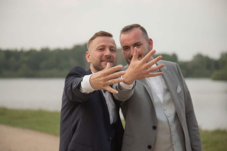 gleichgeschlechtliches Ehepaar, die beiden Männer, zeigen ihre Ringe in die Kamera des Hochzeitsfotografen. Ein tolles Brautpaarshooting zum Sonnenuntergang.